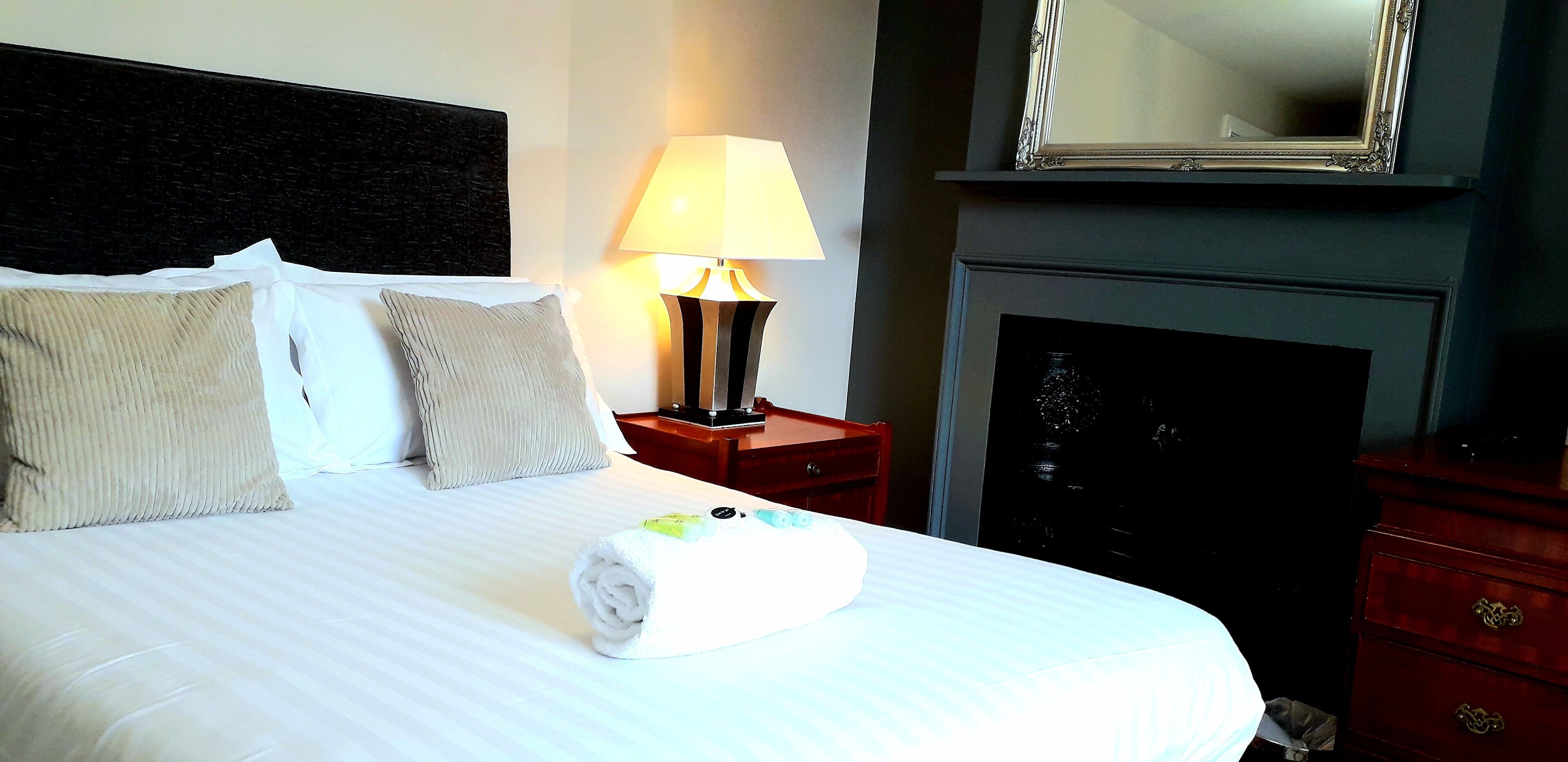 A neatly made bed with pillows, a rolled towel, a bedside lamp, a mirror, and a dresser.