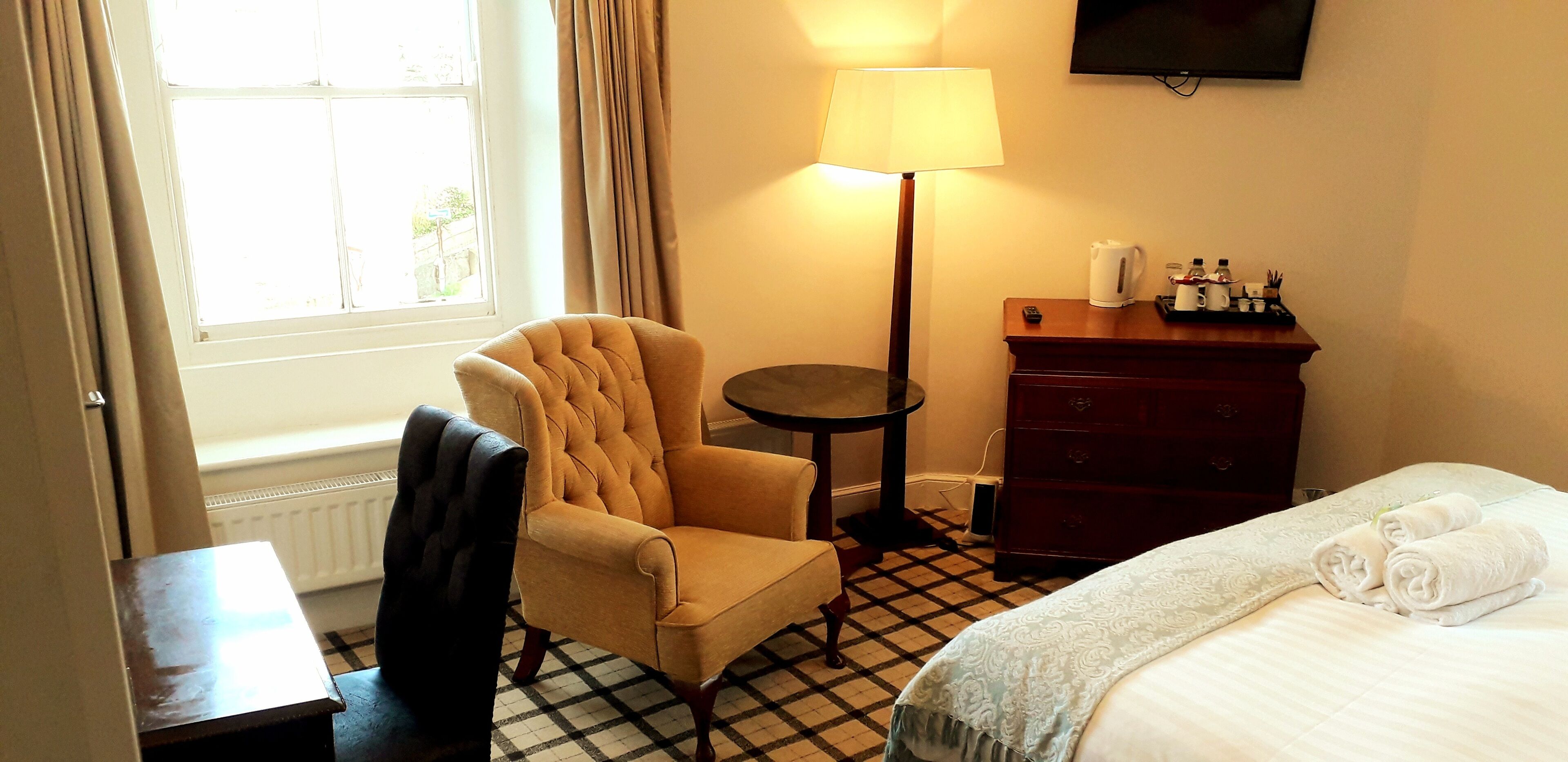 Une chambre d’hôtel avec un sol à carreaux, un fauteuil capitonné, une table de chevet, une lampe, une commode et un lit avec des serviettes.