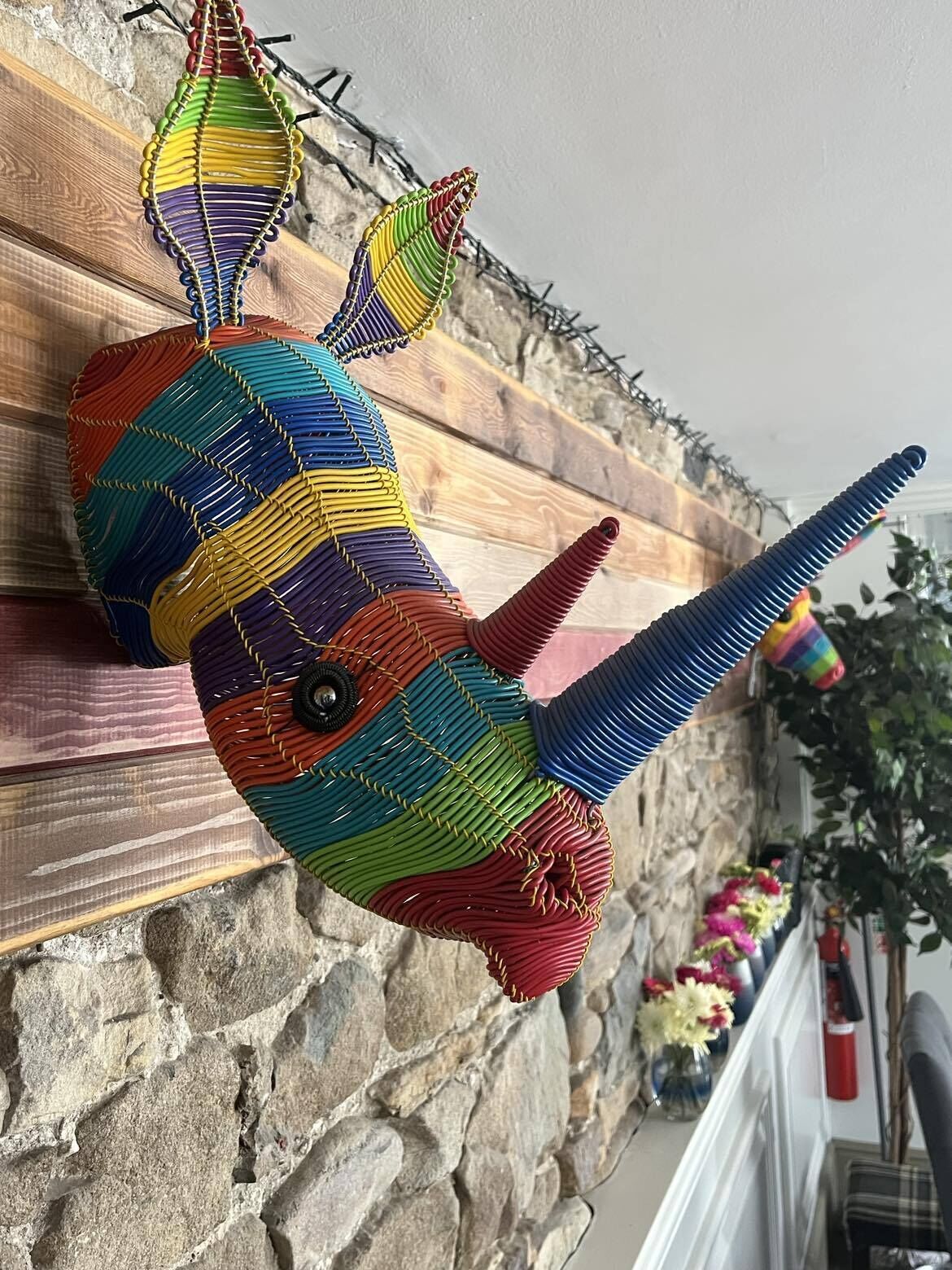 Eine farbenfrohe gewebte Fischskulptur hängt an einer Holzwand.