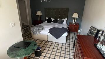 Ein Hotelzimmer mit Schachbrettfußboden, einem Bett mit vielen Kissen, einer Kristallleuchtre, einem Schreibtisch mit einem Laptop und einer Holzkommode.