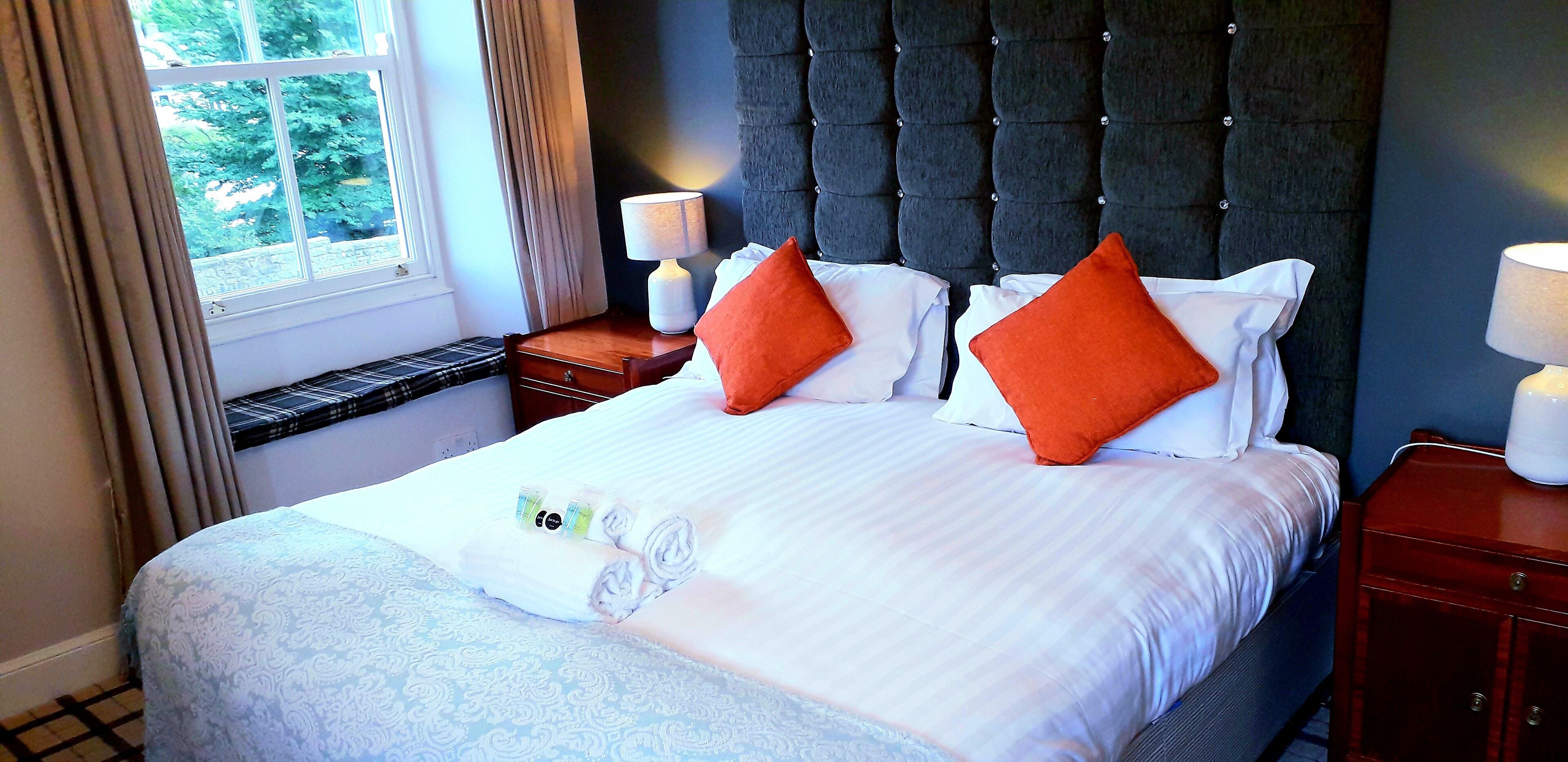 A neatly made bed with white linens and orange pillows, a bedside table with a lamp, and a window with curtains.