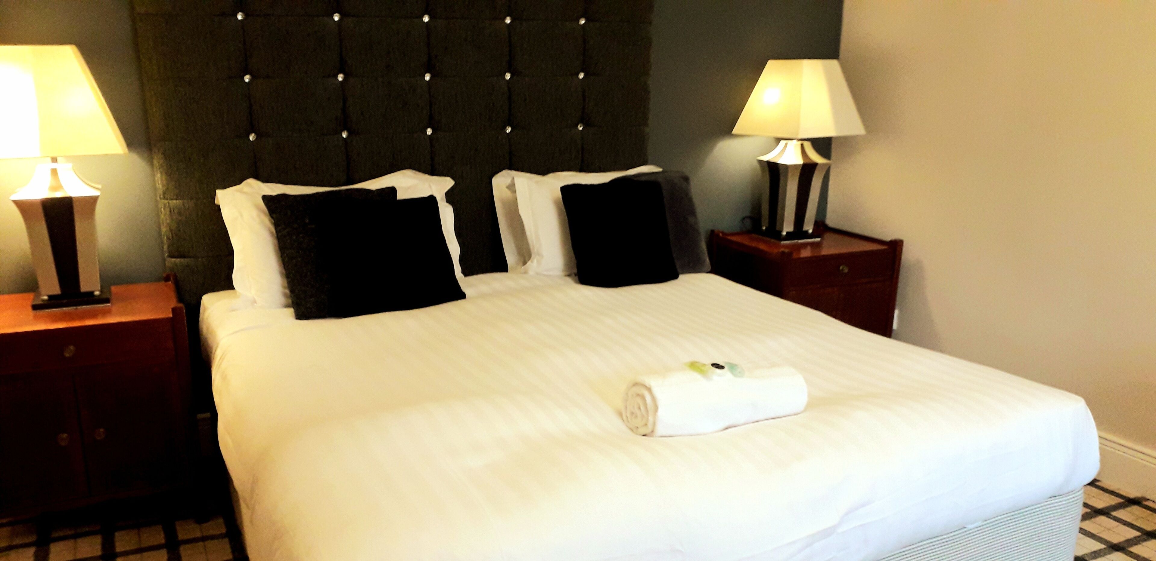 A neatly made bed with white linens, two black pillows, and a rolled towel on the bed. There are two bedside tables with lamps on either side of the bed.