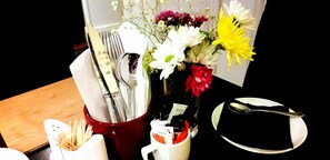 A table set with a red vase containing white and yellow flowers, a white cup with utensils, and a black plate with a spoon. - The Railway Hotel (Hexham)