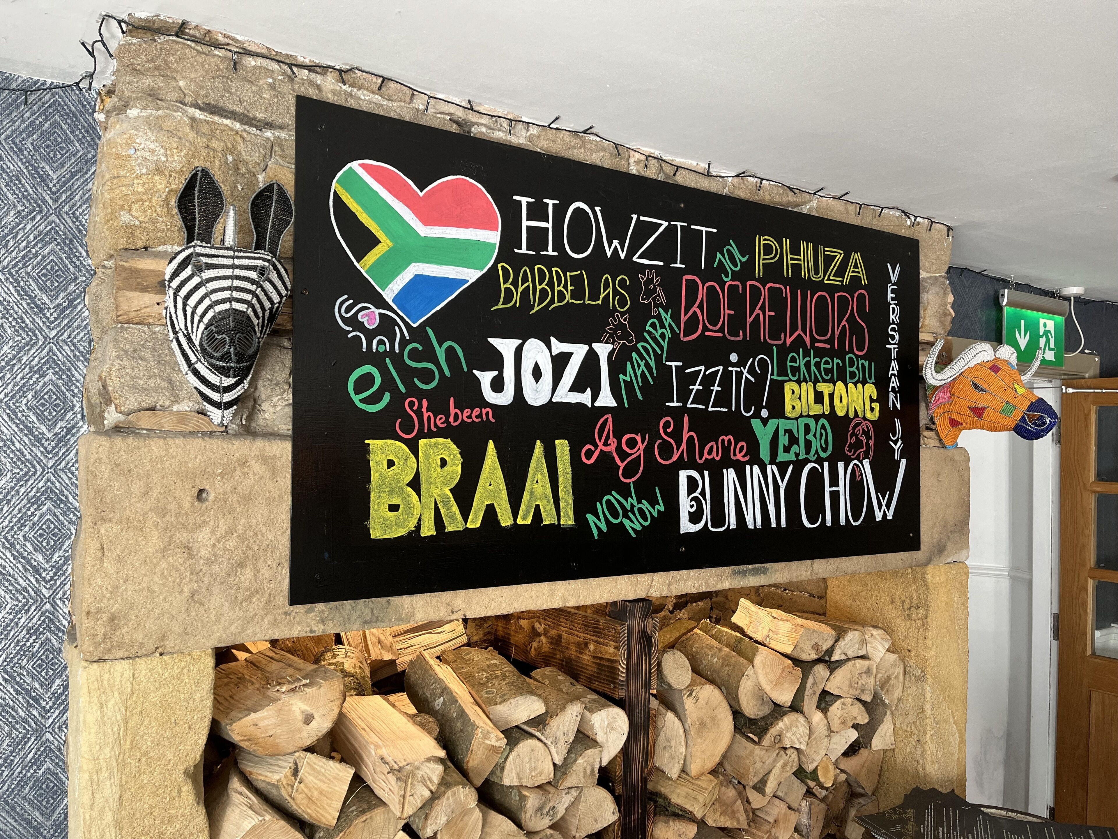 A blackboard with colorful text and a South African flag, surrounded by a rustic stone wall and a pile of firewood.