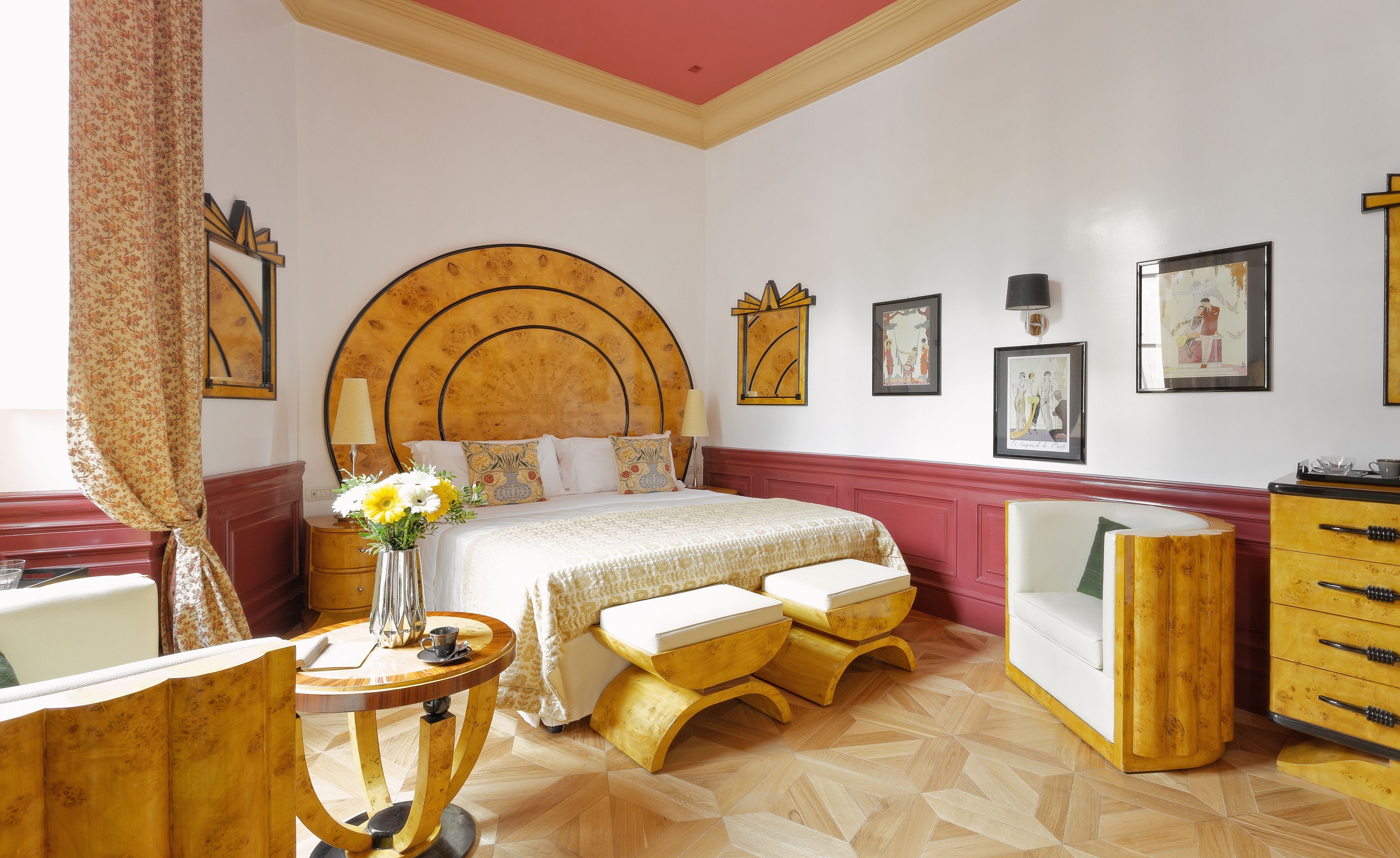 Ein Hotelzimmer mit einem großen Bett, einem hölzernen Kopfteil, einem kleinen runden Tisch mit Blumenvase, einer Holzbank und einer Kommode mit Schubladen.