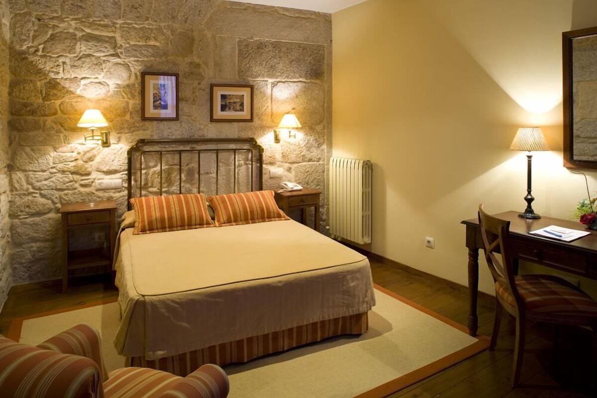 A bedroom with a stone wall, a bed with striped pillows, two bedside lamps, a wooden nightstand, a desk with a chair, and a radiator.
