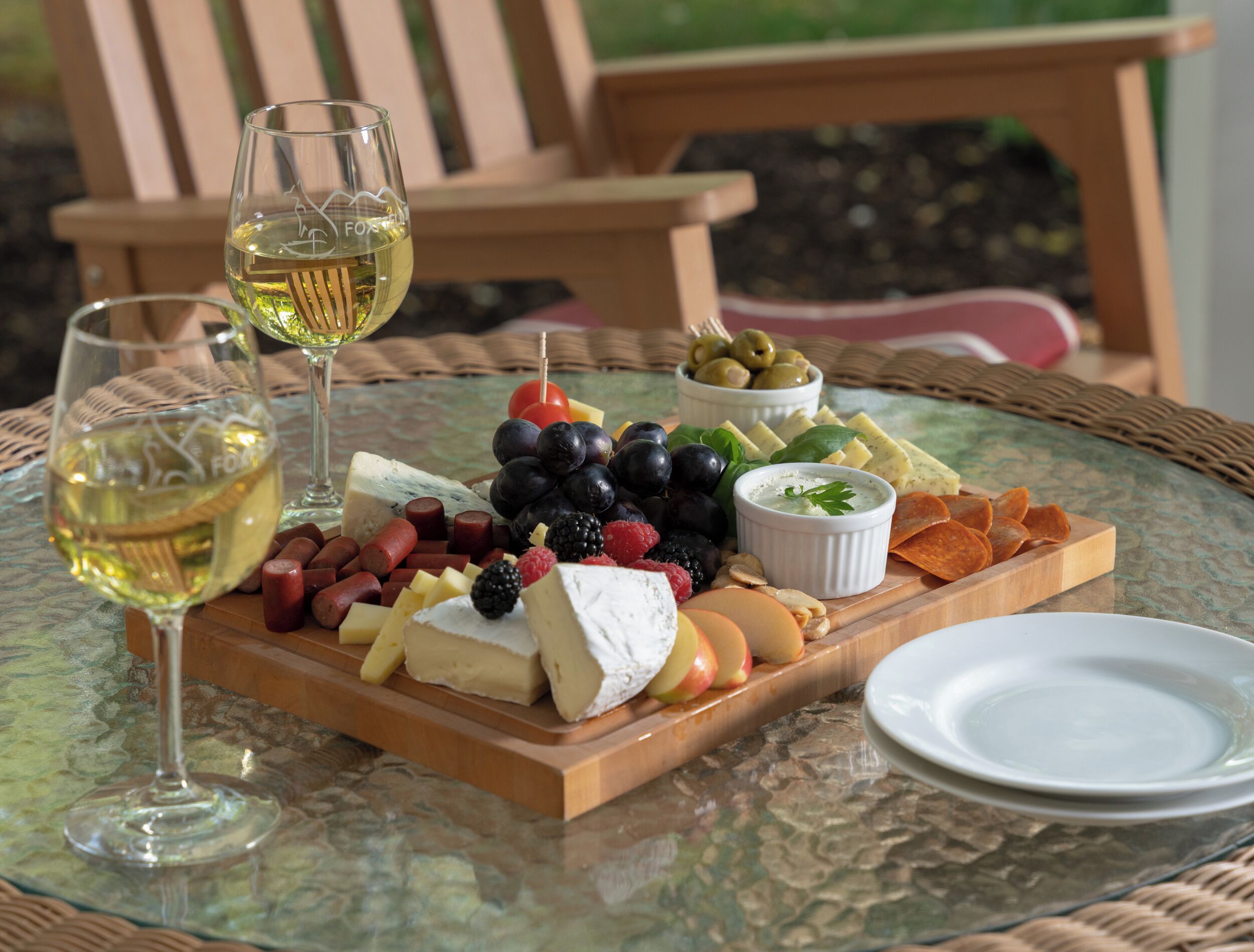 A glass table with a cheese board, grapes, olives, and a bowl of dip. Two glasses of white wine are also visible.