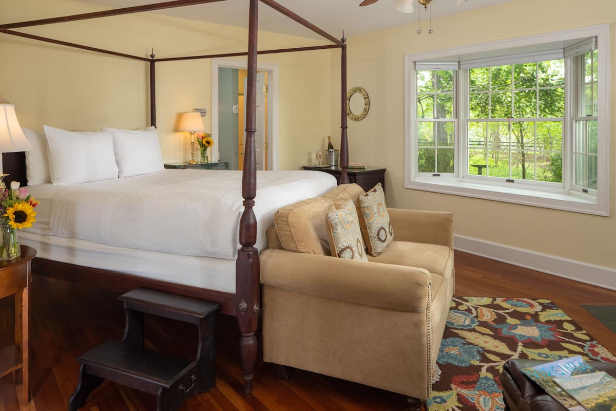 A four-poster bed with a nightstand, a beige armchair, and a window with a view of greenery.