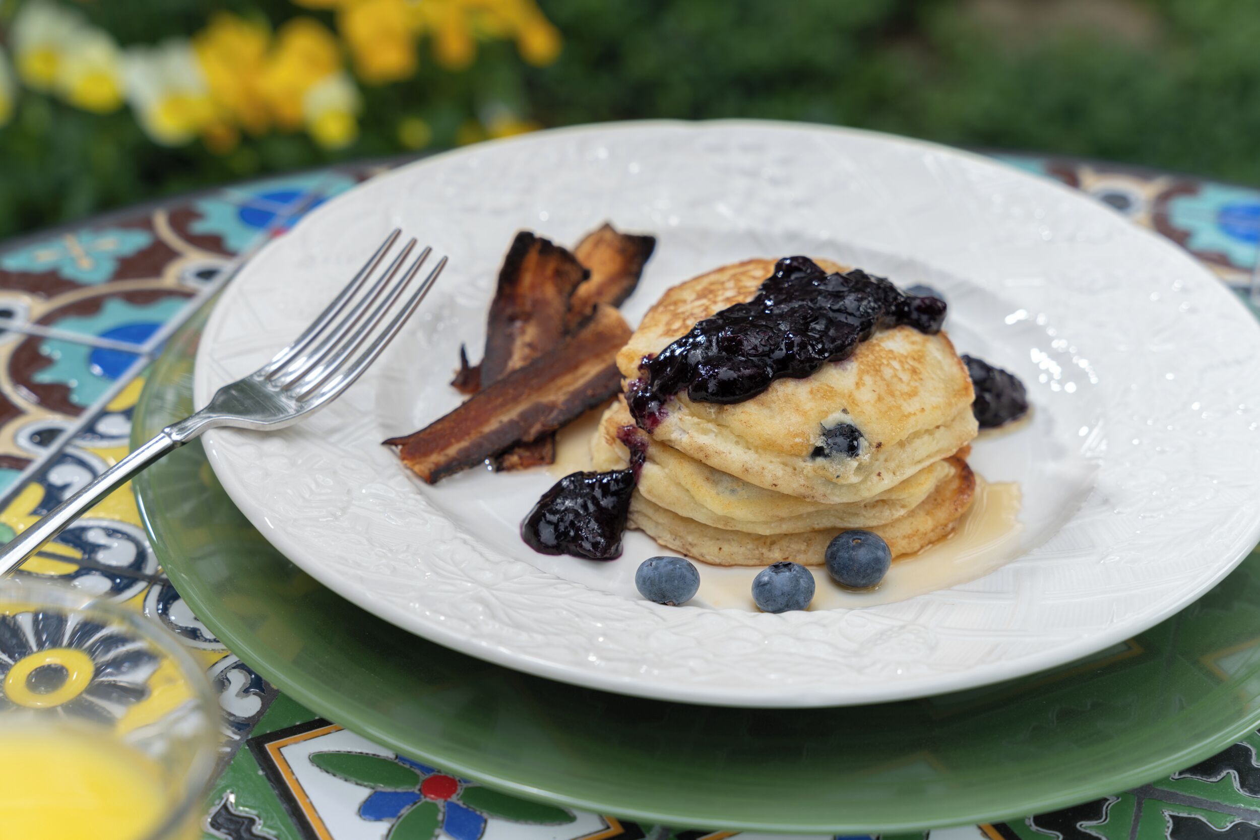 Une assiette de crêpes garnies de myrtilles et nappées de sirop, accompagnée de bacon et d’une fourchette, le tout posé sur une nappe aux couleurs vives.