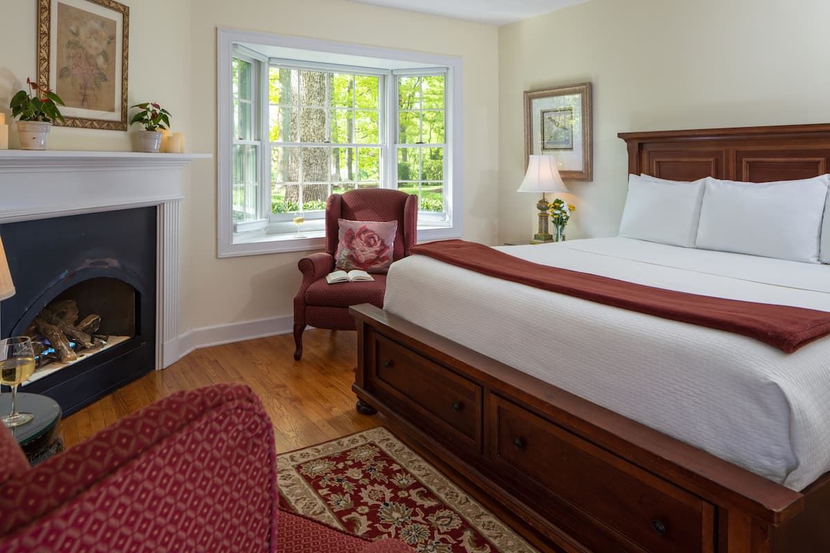 A bedroom with a large bed, a fireplace, a window with a view of trees, and a chair with a book.