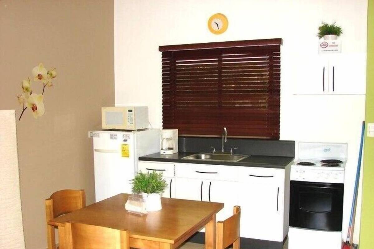 Cocina compacta equipada con microondas, cafetera, lavamanos y estufa. Se observa una mesa de madera con sillas y un reloj en la pared.