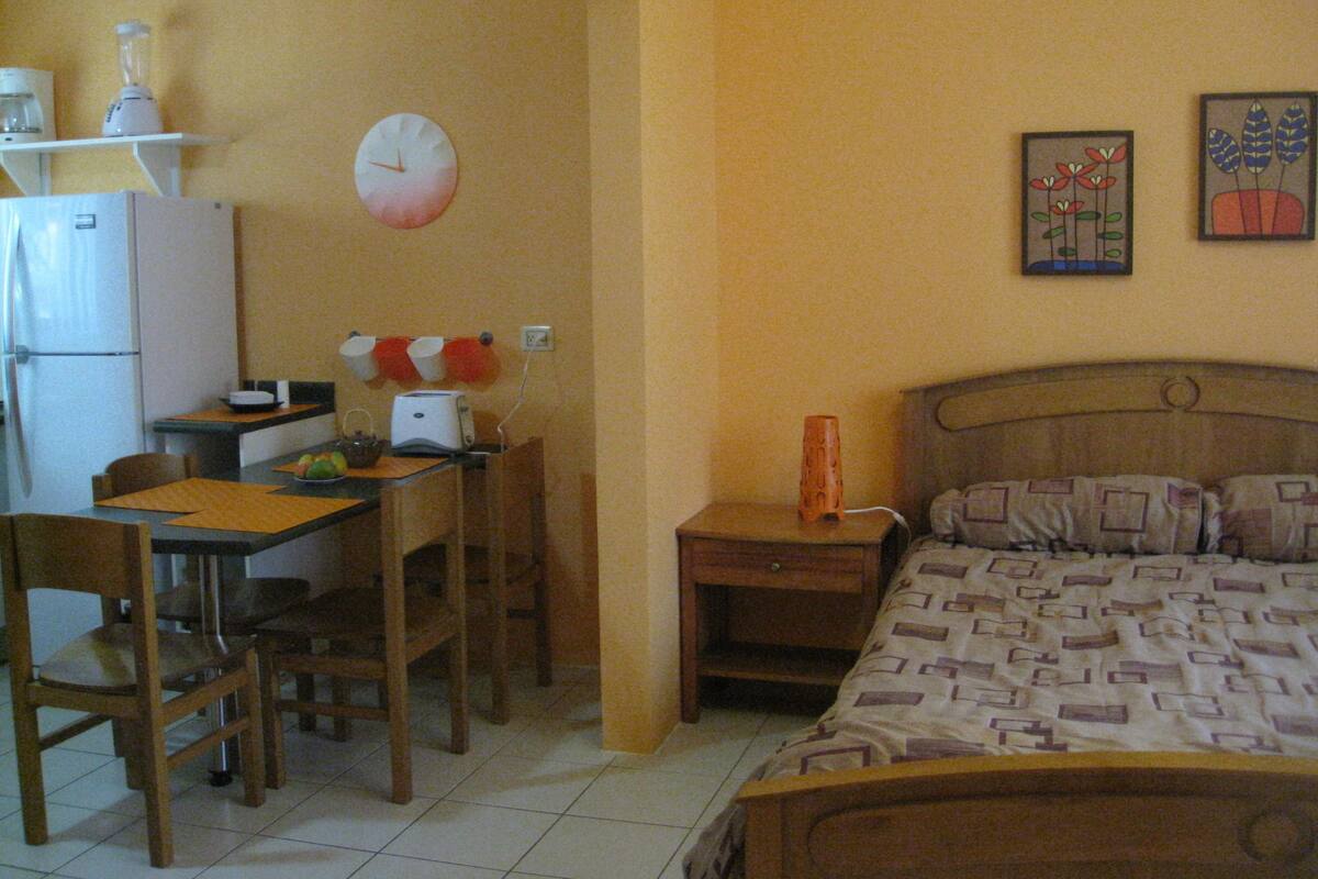 Una habitación pequeña y funcional con cama, escritorio, silla, mesita de noche y un reloj de pared.