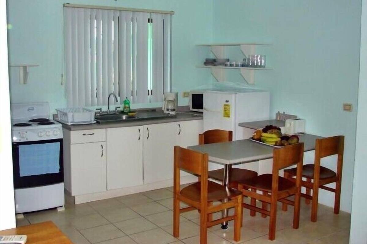Cocina compacta con estufa, lavamanos y refrigerador, una mesa de comedor con sillas y un ventanal con persianas.