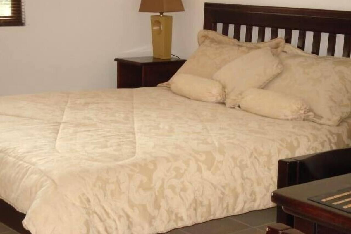 Una cama bien hecha con un edredón beige estampado y dos almohadas, un cabecero de madera y una mesita de noche con una lámpara.