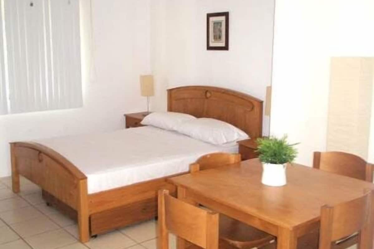 Una cama individual con cabecera de madera, una mesita de noche, una lámpara pequeña, una planta en maceta y una silla de madera.