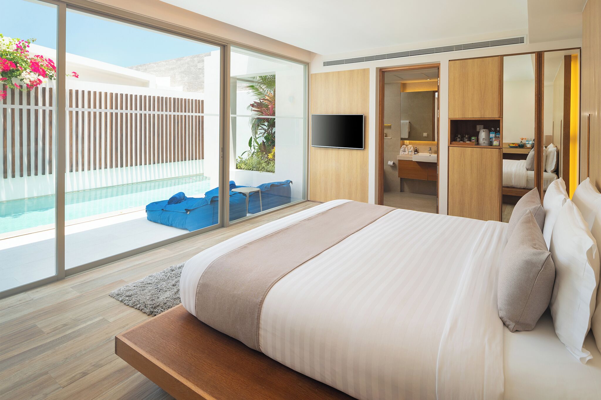 Una habitación de hotel moderna con una cama grande, televisión, vestidor y balcón con piscina y sillas azules.