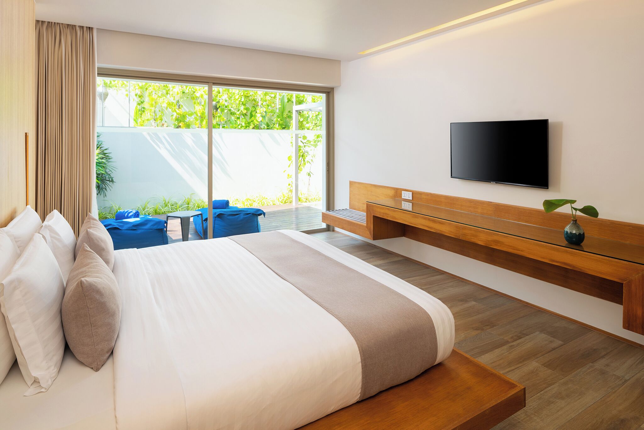 Una habitación de hotel moderna con una cama grande, un televisor montado en la pared, un banco de madera y un ventanal corredero que da a un espacio al aire libre.