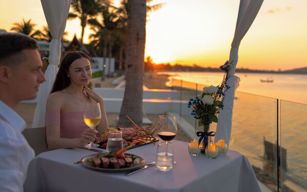 A table set for two with a view of the sunset over water, a glass of wine, and a plate of food.