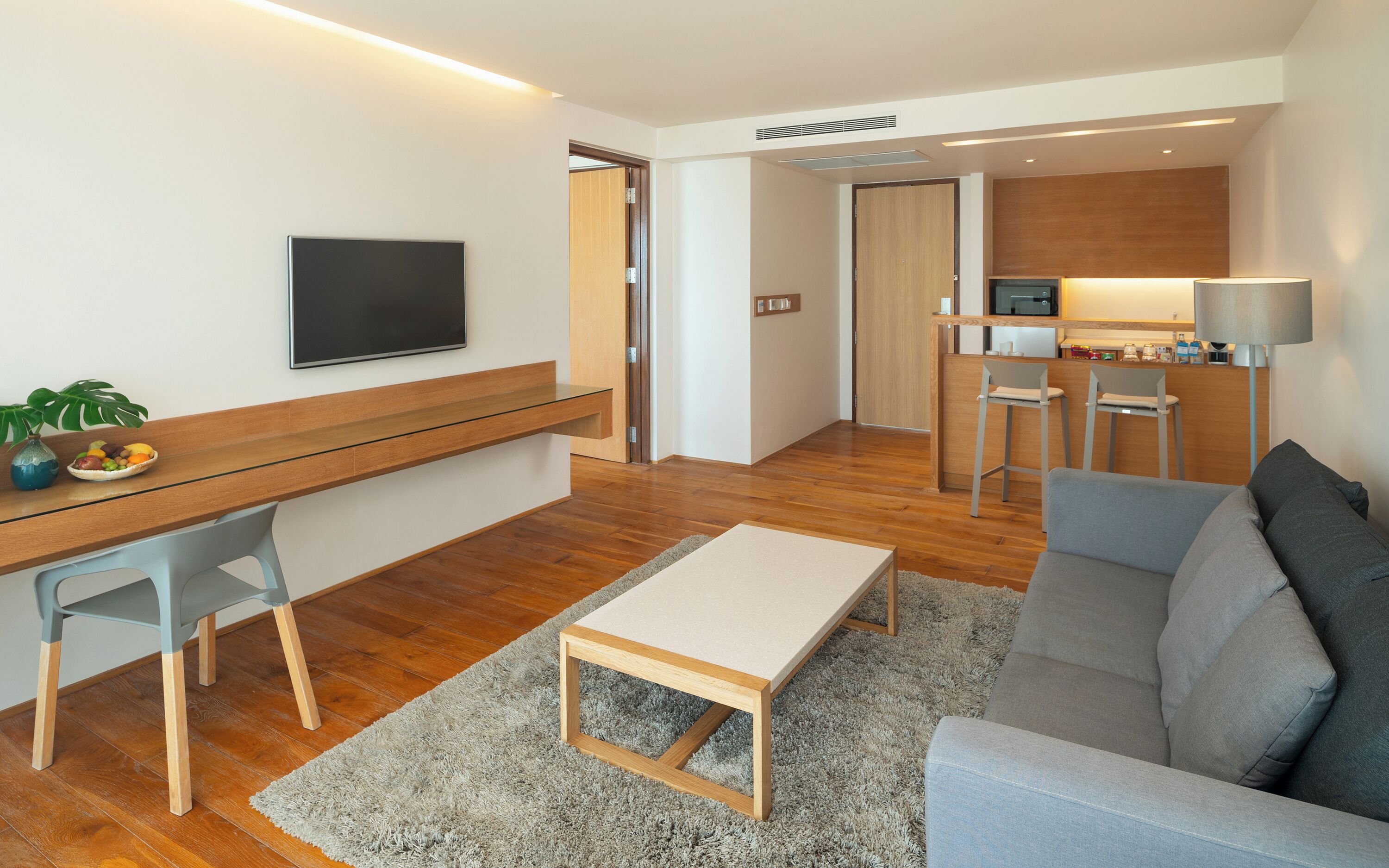 Un salon moderne doté d’un téléviseur à écran plat, d’un bureau en bois, d’un canapé gris et d’un petit coin cuisine avec un four à micro-ondes.
