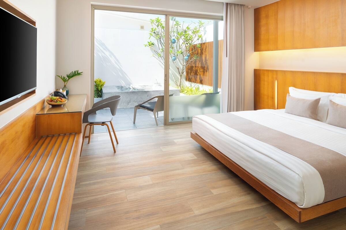 Una habitación de hotel moderna con una cama grande, un televisor de pantalla plana, un escritorio con silla y un balcón con plantas.