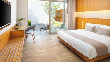A modern hotel room with a large bed, a flat-screen TV, a desk with a chair, and a balcony with plants.
