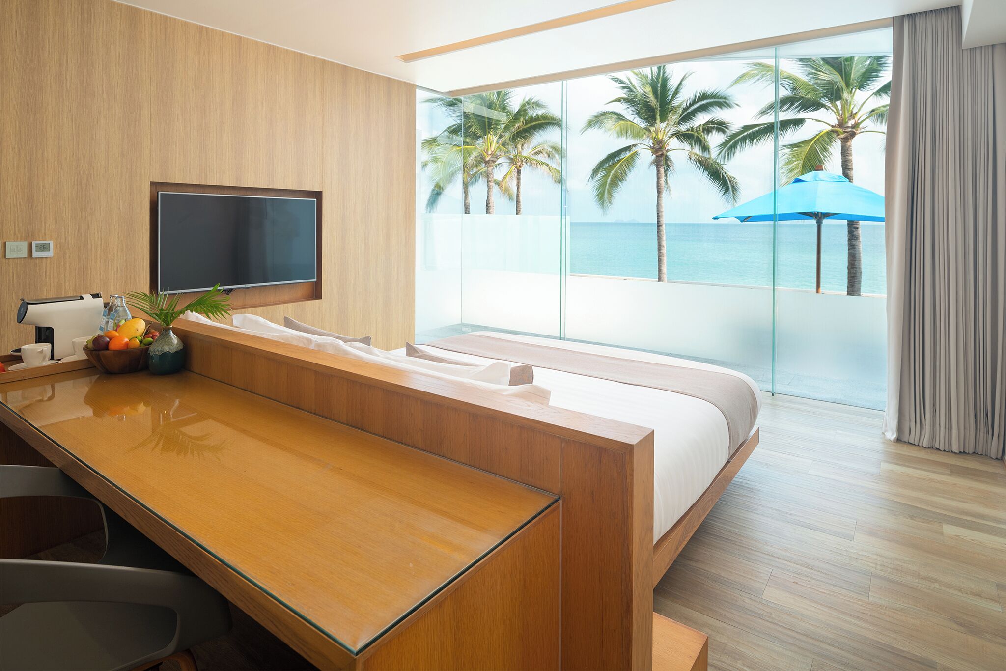 Habitación de hotel moderna con una cama grande, un escritorio de madera, un televisor de pantalla plana y vista al océano con palmeras.