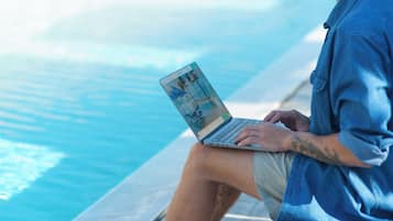 A person sitting by a poolside with a laptop, wearing a denim shirt and shorts.