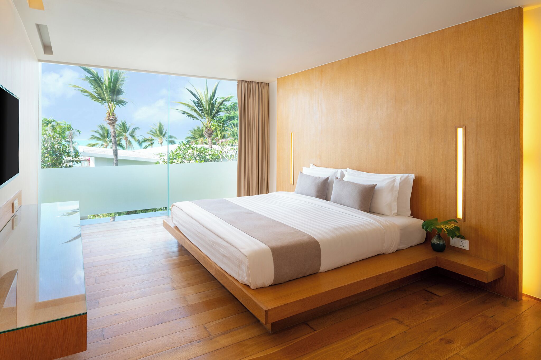 Un dormitorio moderno con una cama grande, televisión, suelo de madera y balcón con vista a palmeras.