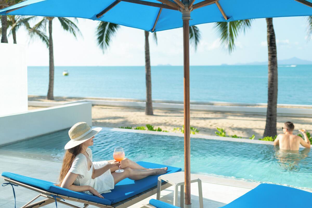 Zona junto a la piscina con sillones y una sombrilla, con vistas a una playa y al océano.