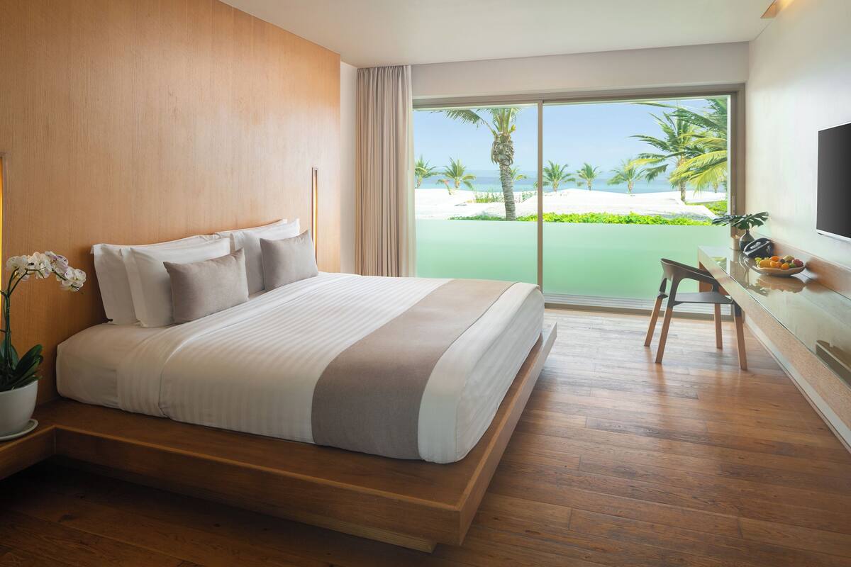 Habitación de hotel con una cama grande, un escritorio con silla, un televisor y vista a la playa.