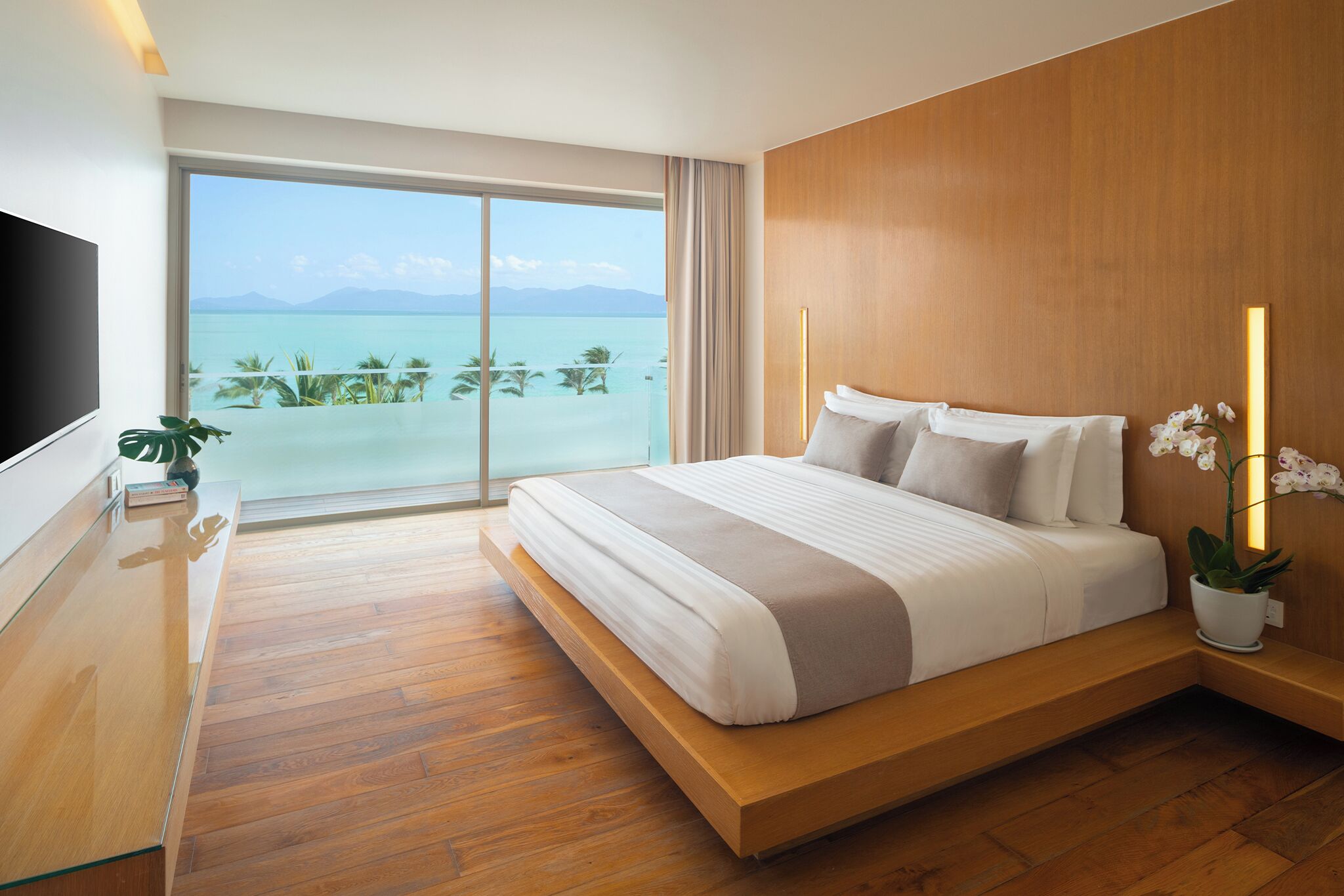 Habitación de hotel moderna con una cama grande, televisión, suelo de madera y vista al mar.