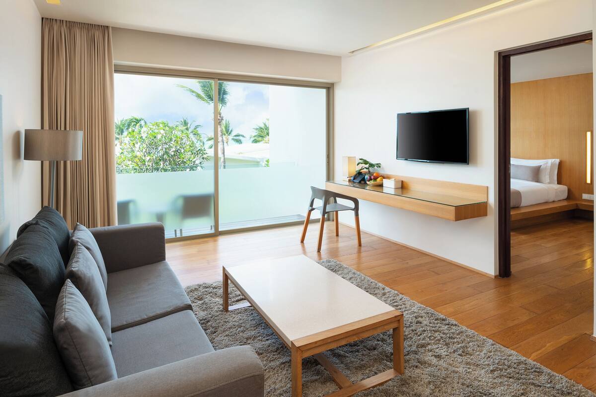 Una habitación de hotel moderna con un sofá, una mesita, un televisor, un escritorio con una planta y un balcón con vistas a palmeras.