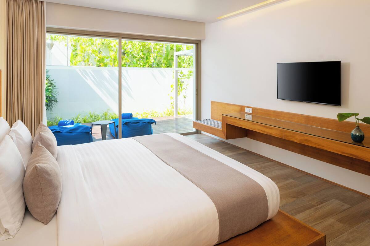 Una habitación de hotel moderna con una cama grande, un televisor montado en la pared, un pequeño rincón de descanso con una mesa y vistas a zonas verdes.