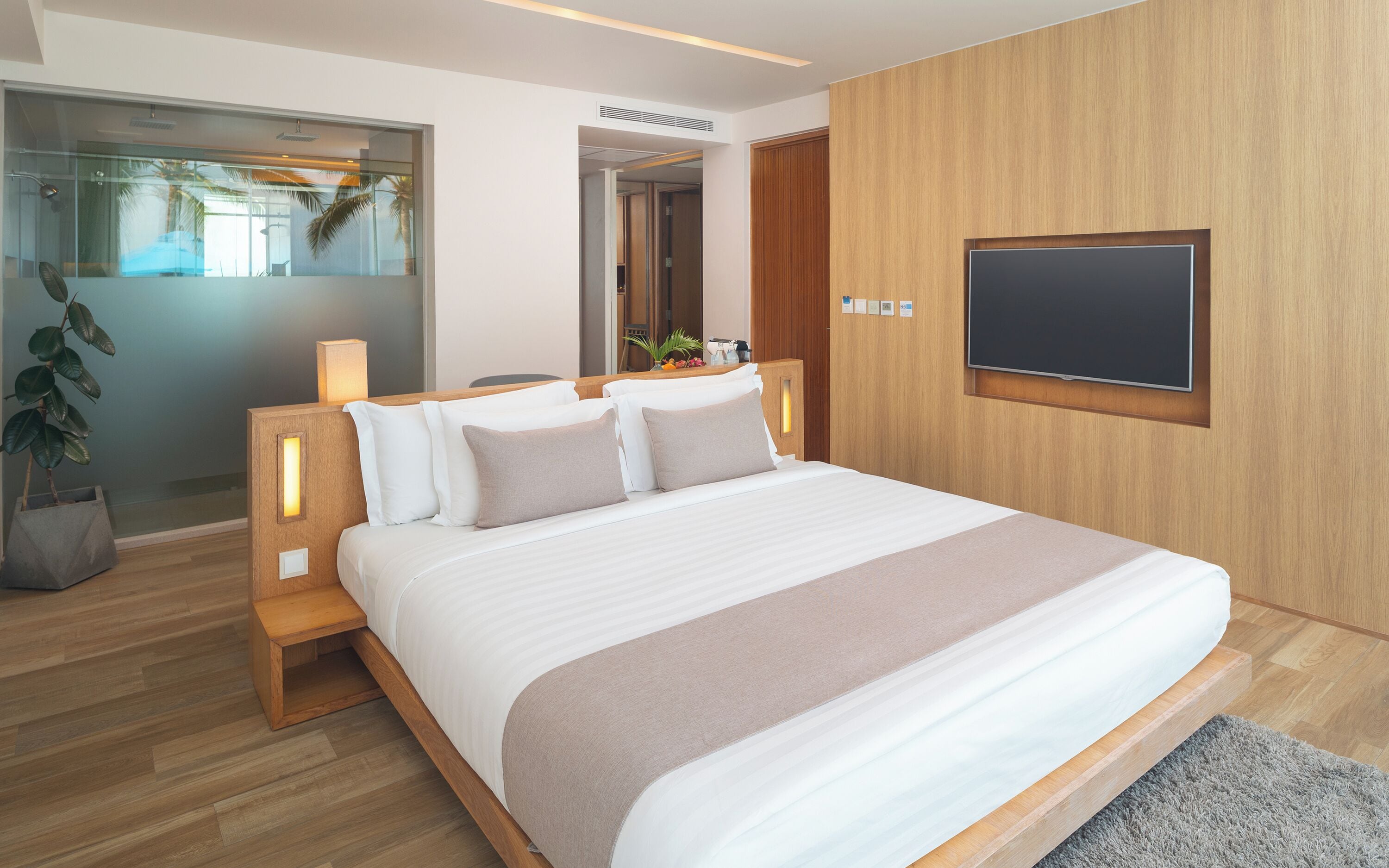 A modern hotel room with a large bed, wooden headboard, bedside lamps, a flat-screen TV mounted on the wall, and a view of palm trees through the window.