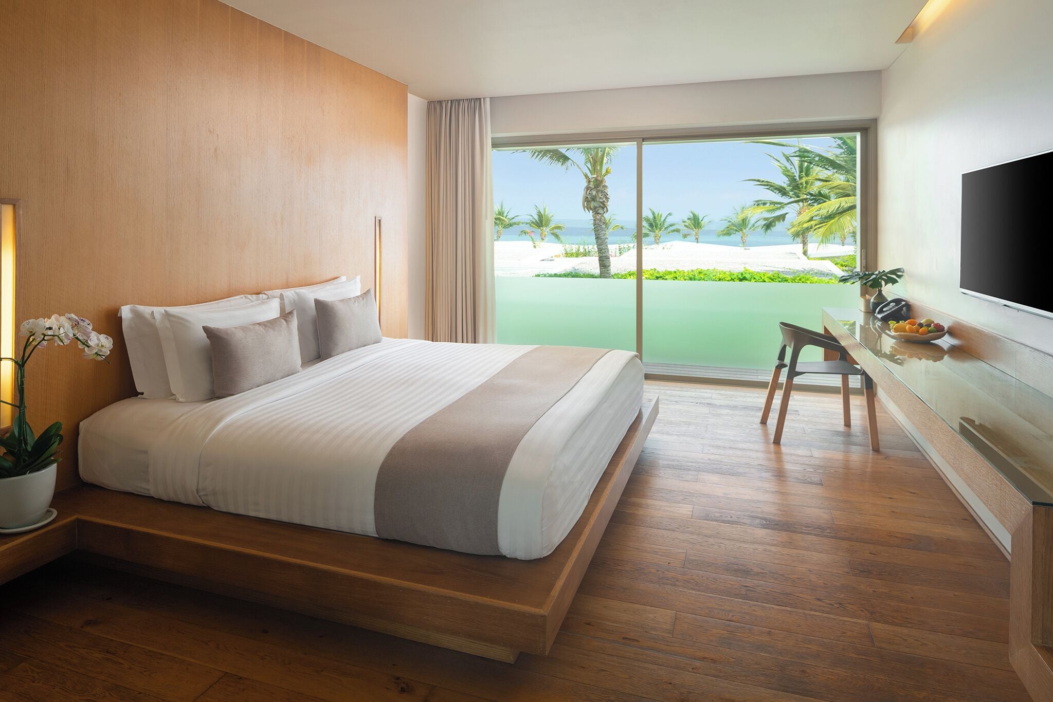 Habitación de hotel moderna con una cama grande, un escritorio, un televisor y vista a la playa.