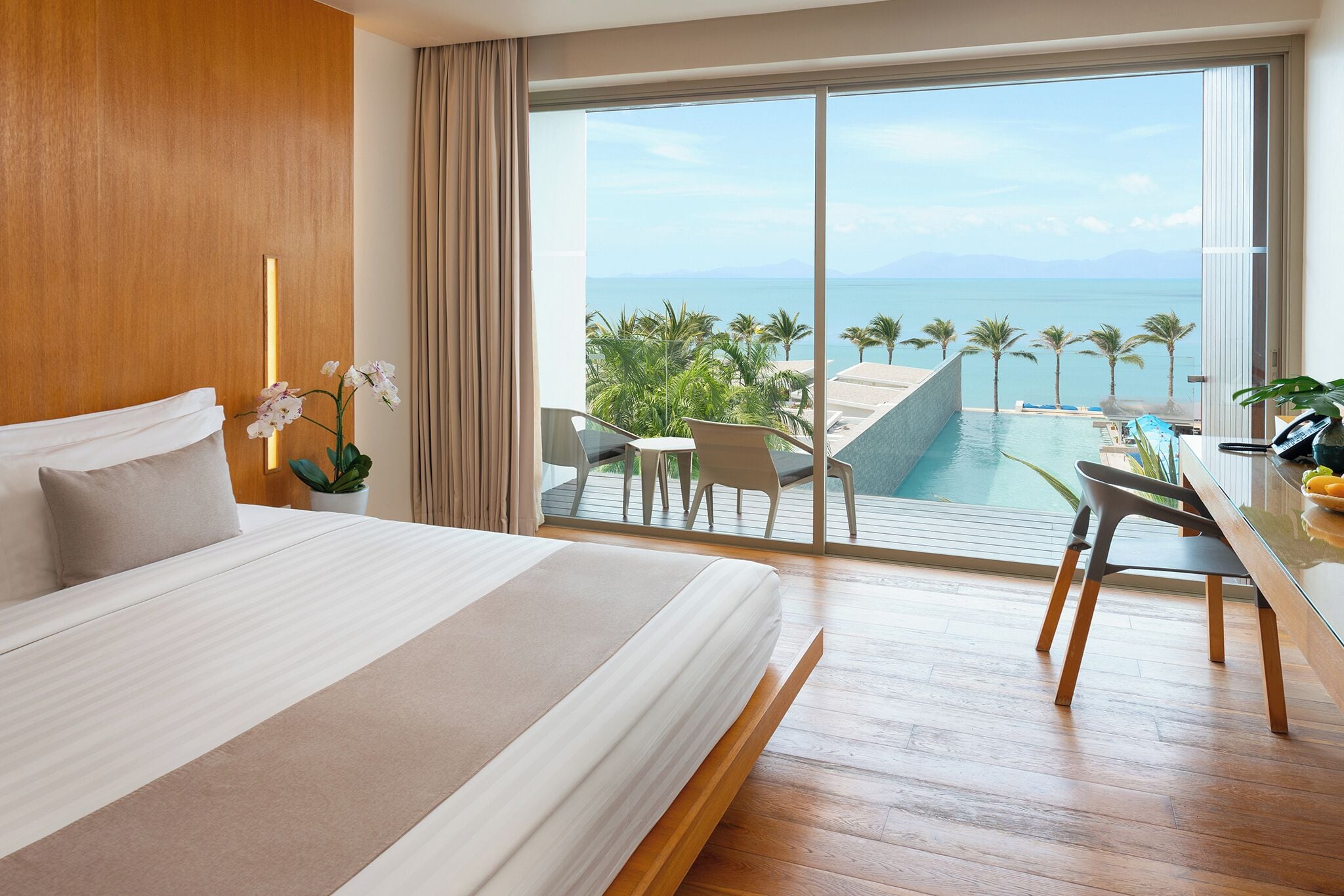 Habitación de hotel con una cama grande, un escritorio con silla, vista al mar y zona de piscina.
