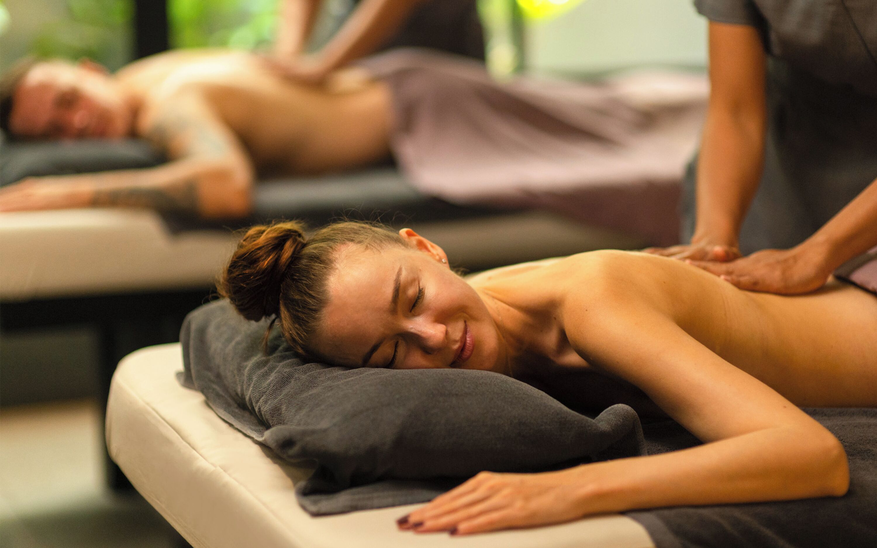 Ein Massageräum mit einer Person, die auf einer Massageliege liegt, einer weiteren Person, die sich um den Klienten kümmert, und einer dritten Person im Hintergrund.