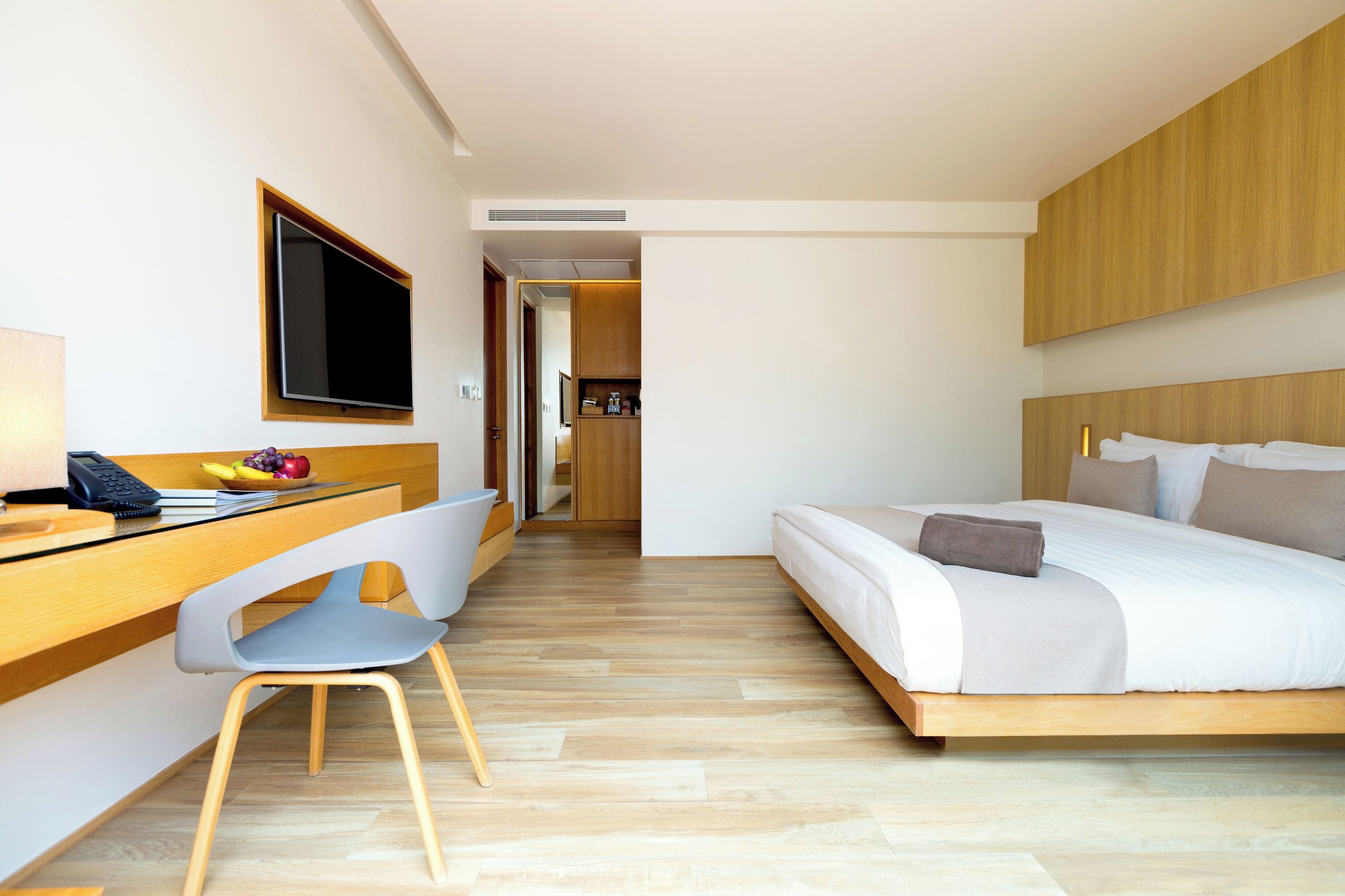 A modern hotel room with a wooden desk, a flat-screen TV, a bed with a wooden headboard, and a small kitchenette in the background.