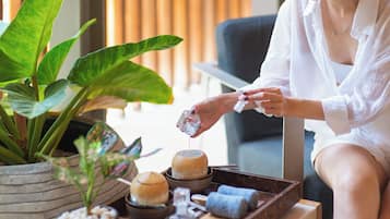 Une personne assise près d’une fenêtre, avec un plateau contenant des articles de toilette, une plante en pot et une table en bois.