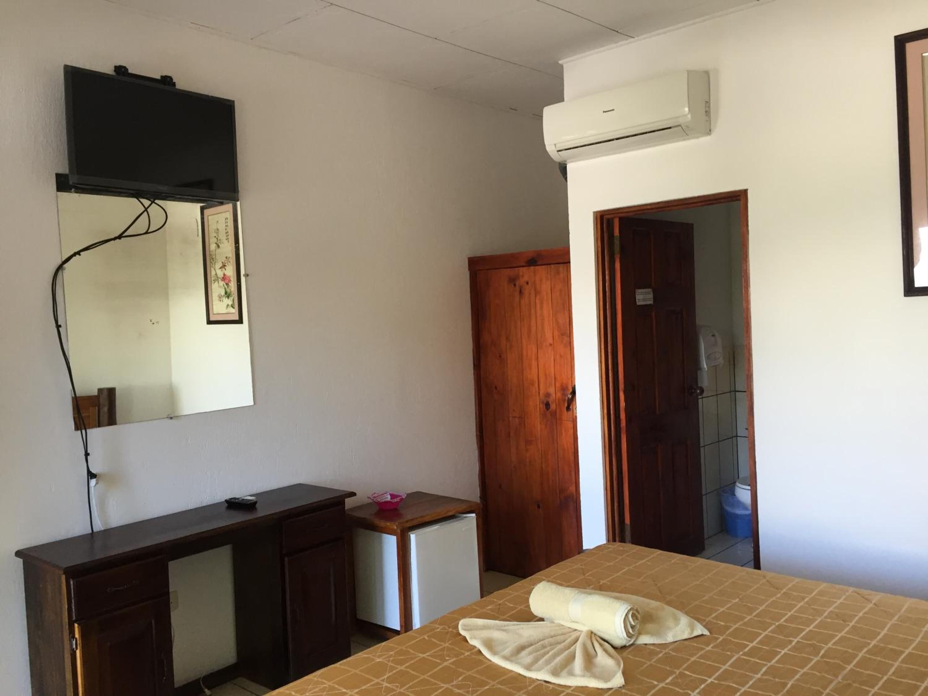 A room with a bed, a wooden desk, a small refrigerator, a wardrobe, and an air conditioning unit.