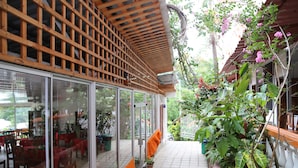 Un espacio al aire libre techado con mesas y sillas, rodeado de plantas en macetas y una estructura de madera celosía.