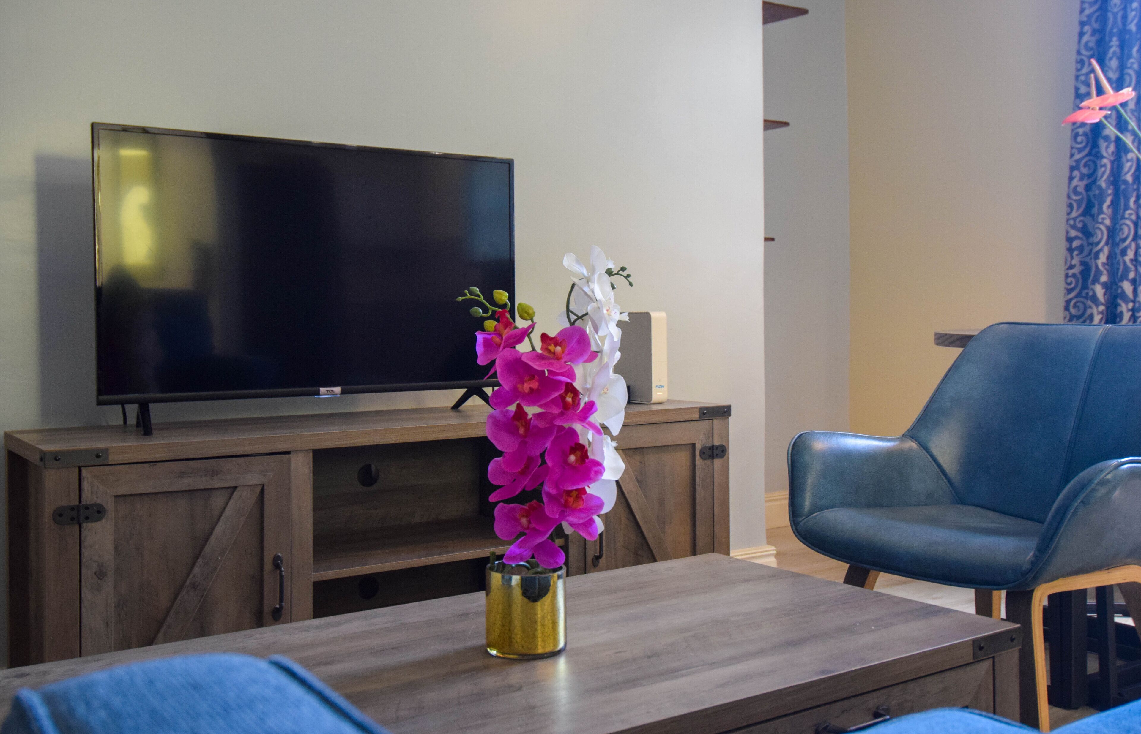 Una sala de estar con un televisor de pantalla plana sobre un mueble de madera, un sillón de cuero azul y una mesa con un jarrón de orquídeas.