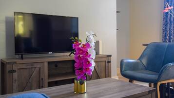 Una sala de estar con un televisor de pantalla plana sobre un mueble de madera, un sillón de cuero azul y una mesa con un jarrón de orquídeas.