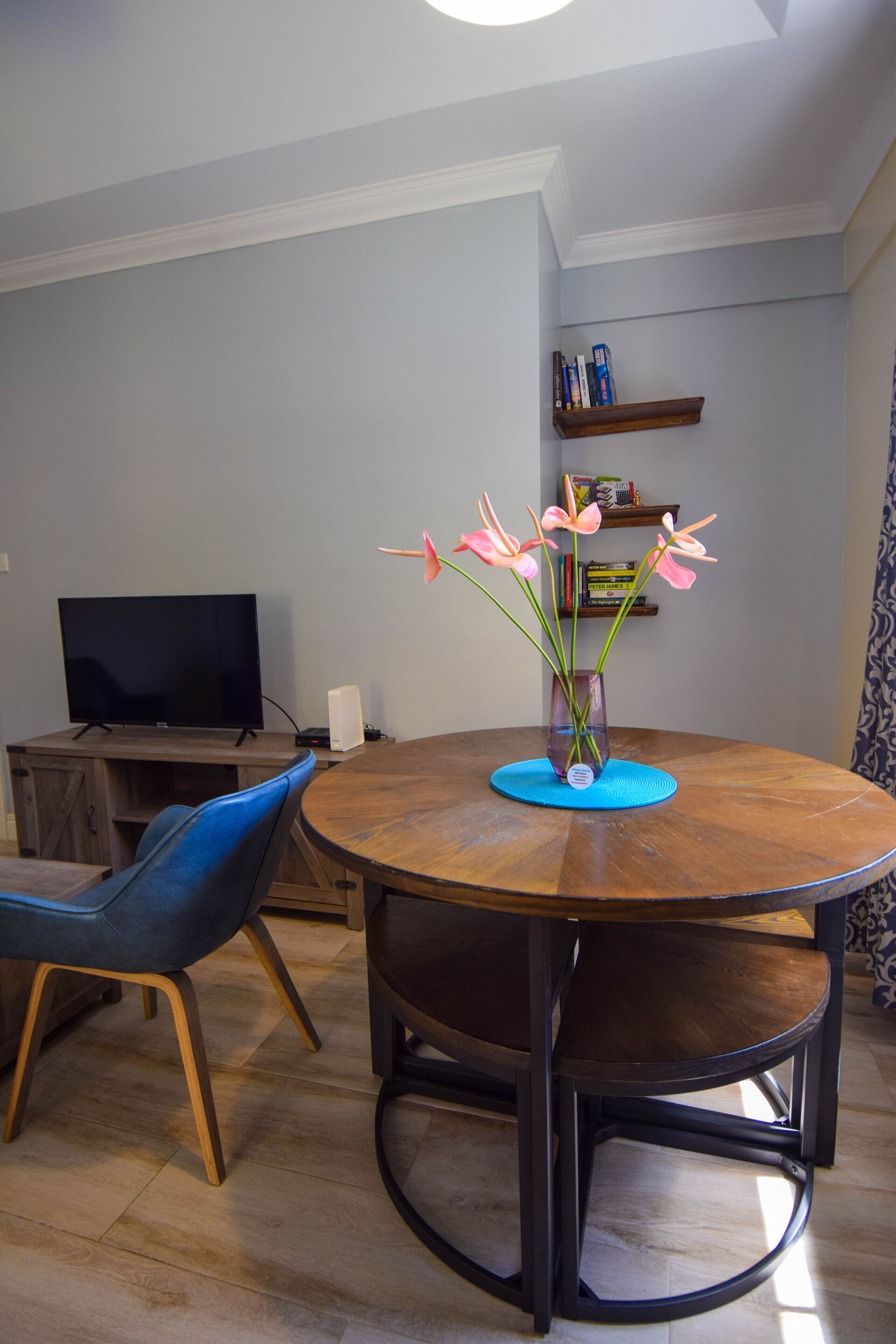 Een eetruimte met een ronde houten tafel, een vaas met bloemen, een blauwe stoel en een televisie op een meubel.