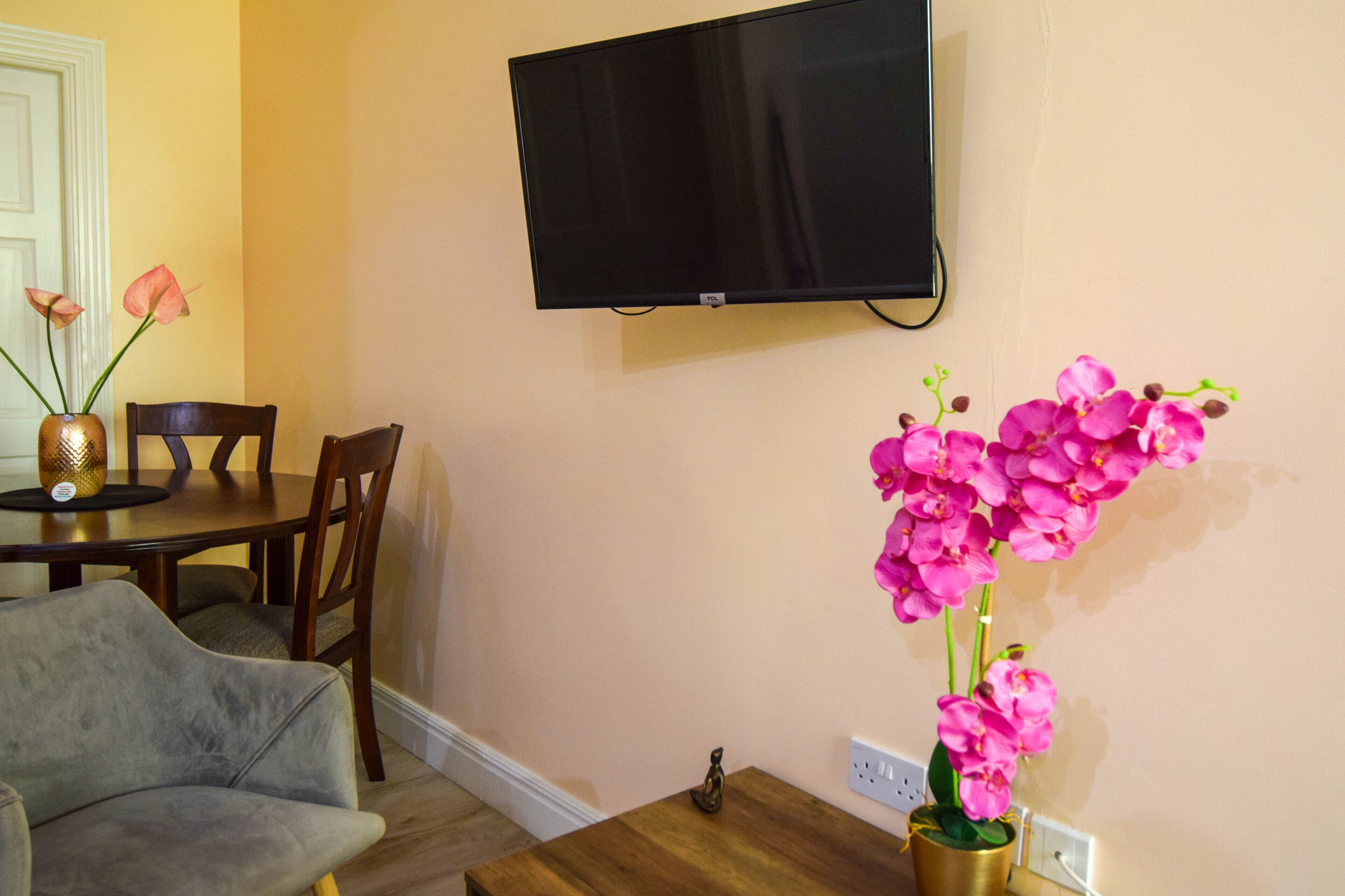 A dining area with a table, chairs, a television mounted on the wall, and a vase with pink flowers.