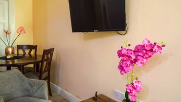 A dining area with a table, chairs, a television mounted on the wall, and a vase with pink flowers.