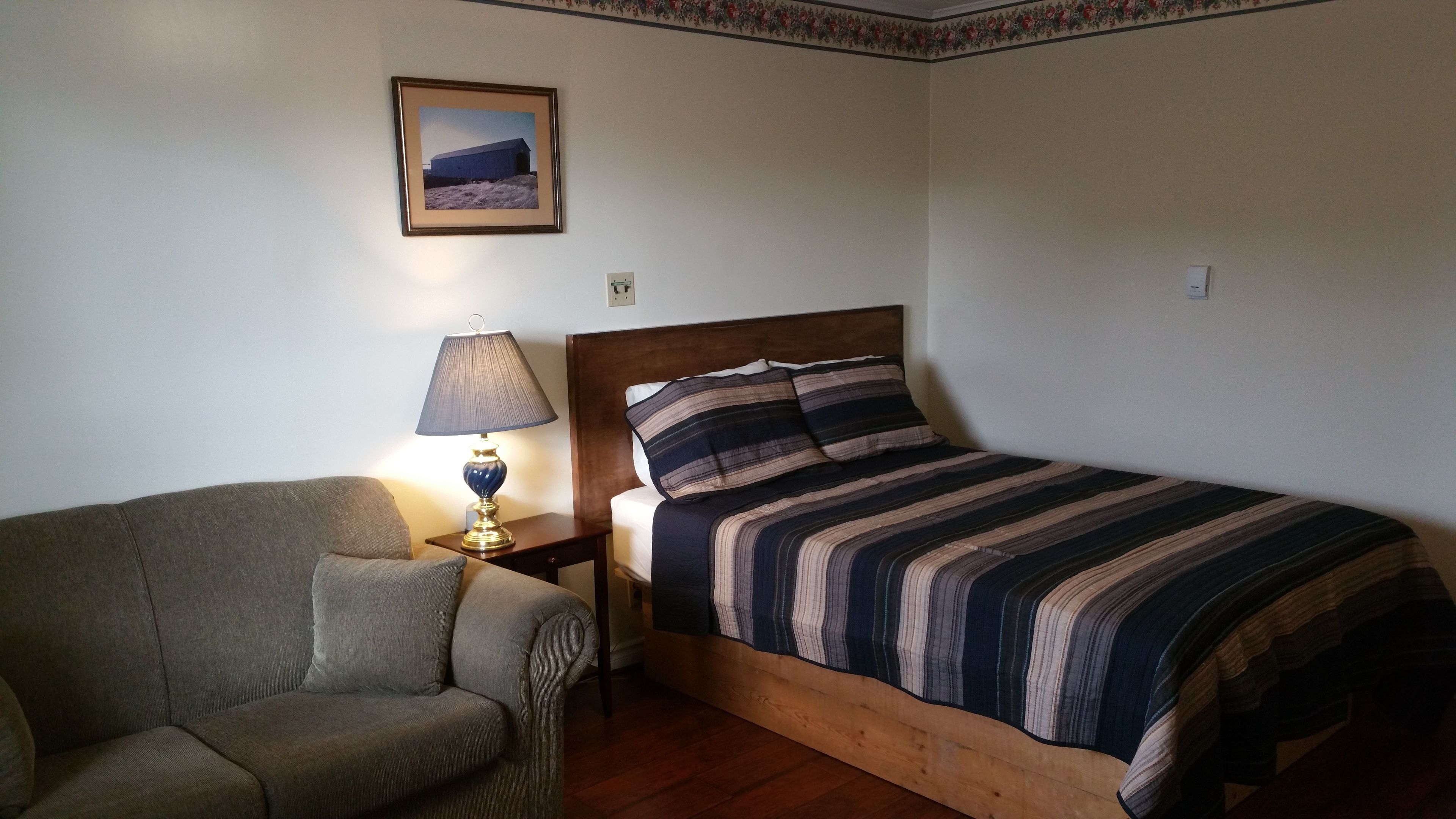 Un dormitorio con cama, mesita de noche, lámpara, una silla y un cuadro en la pared.