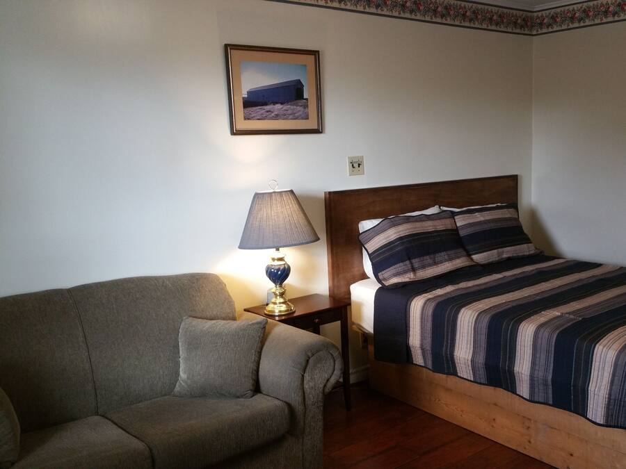 Un dormitorio con cama, mesita de noche, lámpara, una silla y un cuadro en la pared.