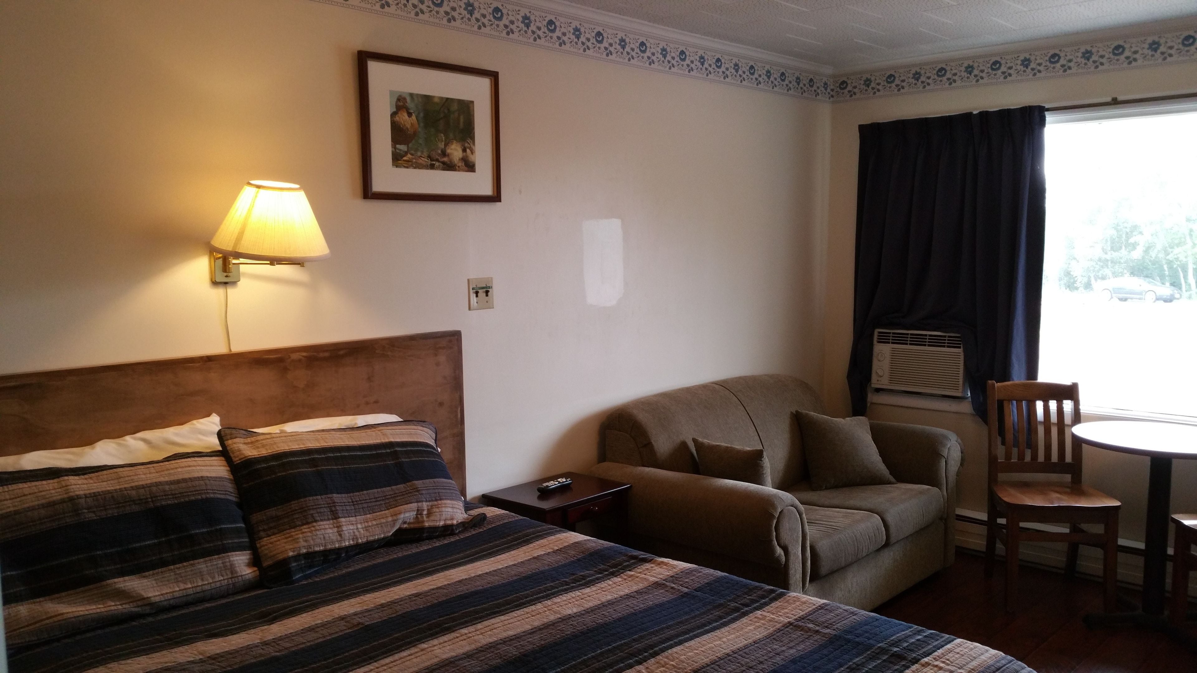 Habitación de hotel con cama, sofá, una mesita y una silla. Hay una lámpara en la pared y un marco de fotos sobre el sofá.