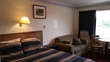 Habitación de hotel con cama, sofá, una mesita y una silla. Hay una lámpara en la pared y un marco de fotos sobre el sofá.